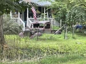 Old rusting truck in front yard under American flag