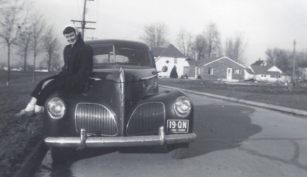 Woman sitting on a 1941 Ford on a road in black & white