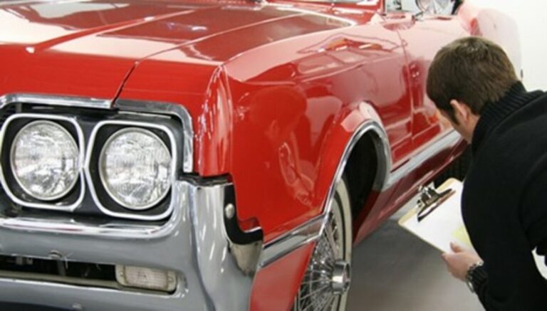 A man with a clipboard inspecting the left side of a 60s red Oldsmobile