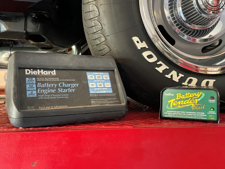 A battery charger and trickle charger on a red car lift next to a front tire
