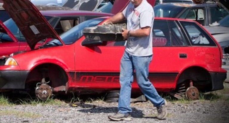 A man iin jeans carrying a fuel tank walking in front on a red Chevy Metro in a junkyard