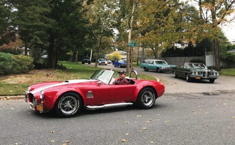 A red with white stripes Cobra driving thru a residential neighborhood with other classic cars turning behind it