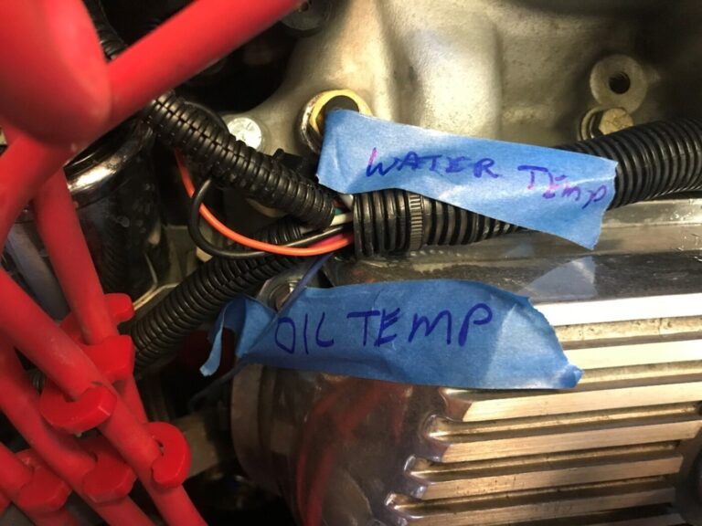 Engine wiring labeled with blue painter's tape and blue marker noting the water temp wire and the oil temp wire on a Ford engine prior to disassembly for restoration planning