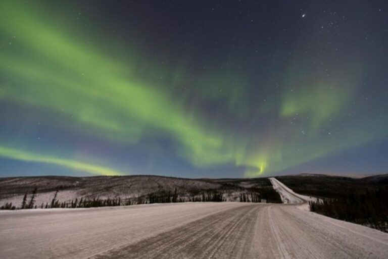 A view of the green shining Northern Lights in blue night sky over the Dalton Hwy Alaska with the snow covered curvy road