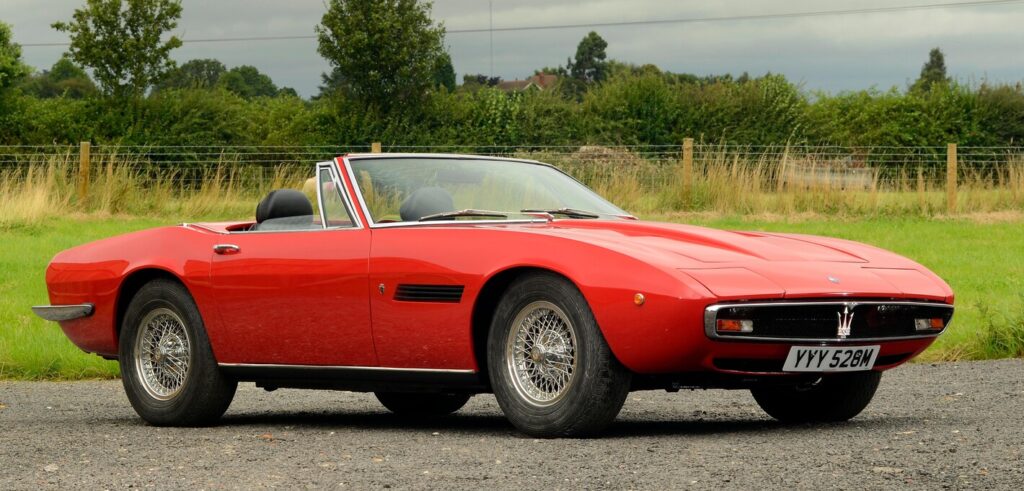 A red 1971 Maserati Ghibli convertible parked in a lot next to grass; is it real or a counterfeiter scams