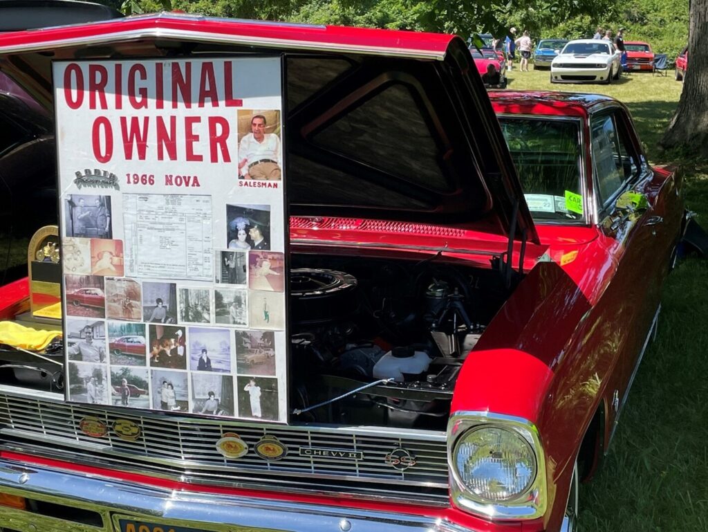 A red 1966 Chevy Nova at a classic car show with ownership sign wit hood open after classic car show display preparation