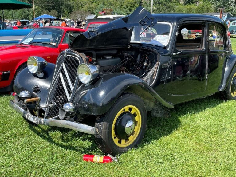 A black Citroen with yellow wheels with its hood open at a car show with a red fire extinguisher on green grass that was ready for car show display preparation