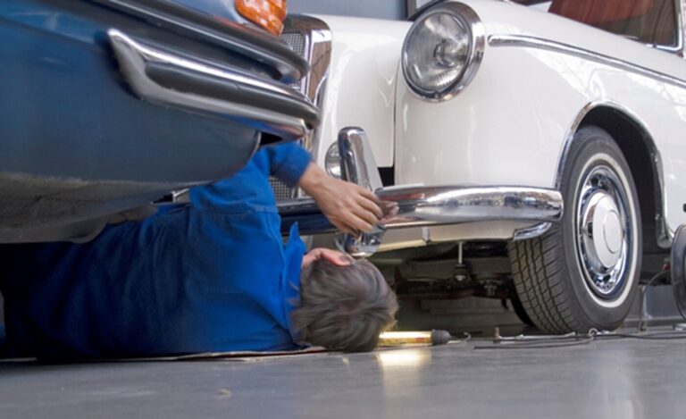 A mechanic in a blue short inspecting under the front of a white Mecedes before starting any autobody repair