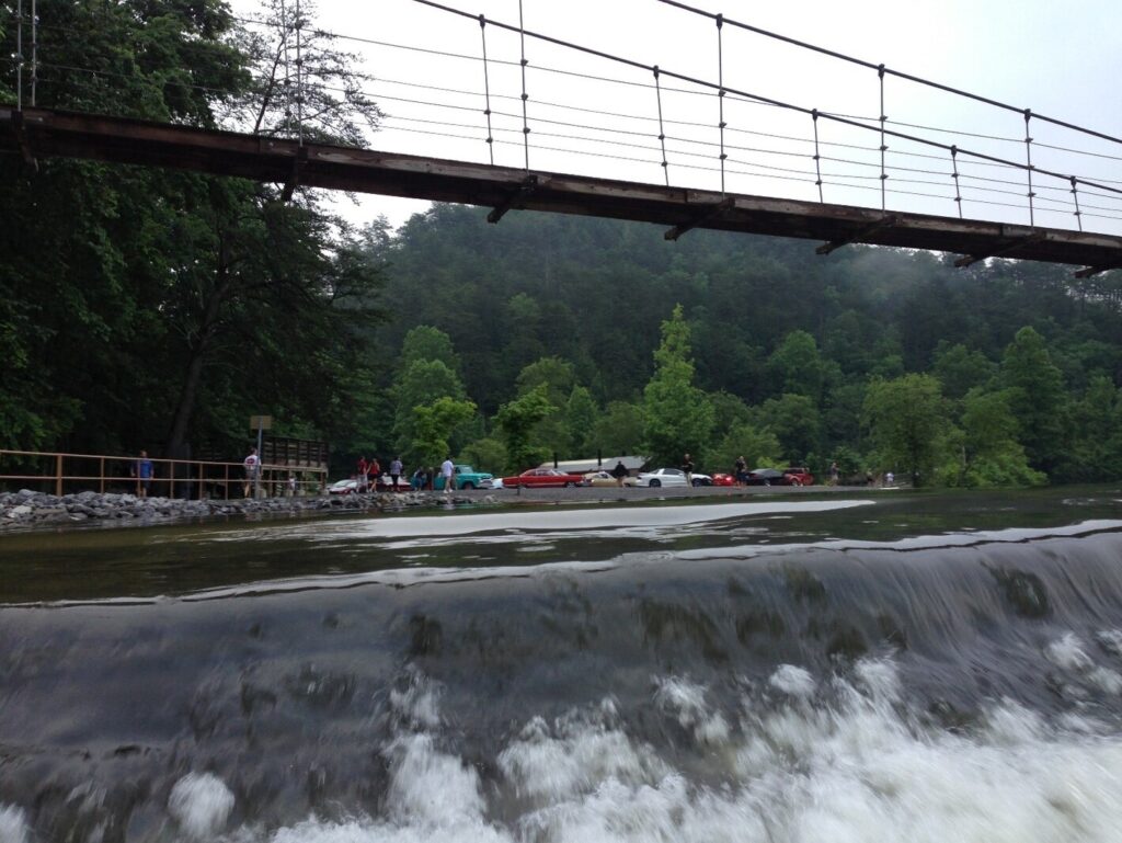 A suspension walking bridge over a water fall with classic cars in the background for waterside road trips