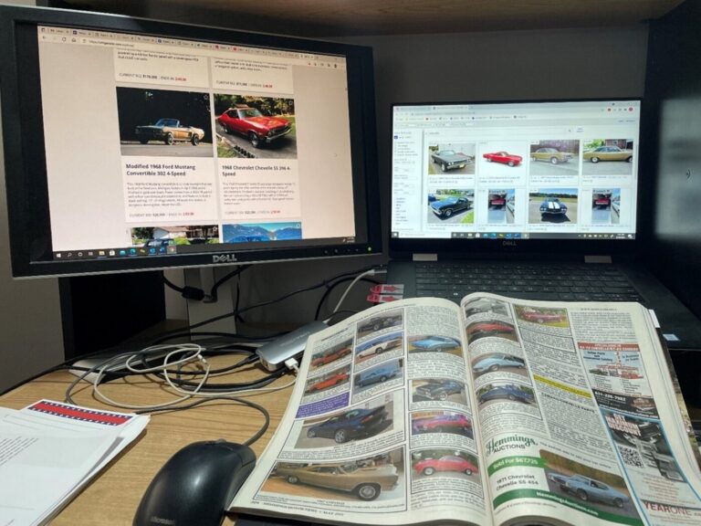 A desk with 2 computer screens showing classic cars for sale and a Hemmings magazine research cars to buy before starting a car restoration