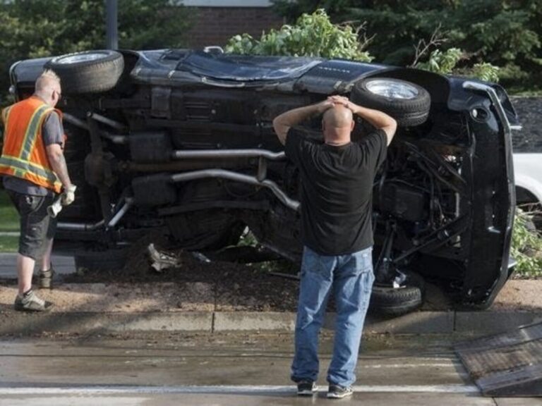A Ford Mustang resting on its side after an accident with the drive in a black shirt and blue jeans looking at the car with his hands on his head and a worker with safety vest on assessing the damage show the need for classic car insurance