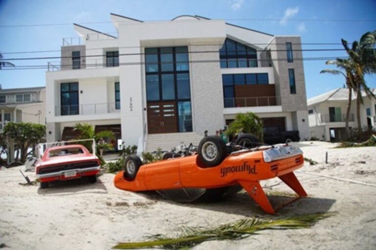 An upside down orange Plymouth Super-bird and a red Dodge Daytona both sitting in the sand damaged in front of a 3-story house showing the need for classic car insurance