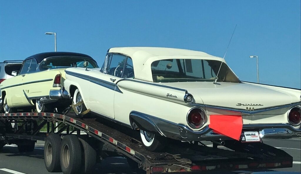 An open car trailer with 2 yellow cars with the rear Ford Fairlane convertible with an orange warning flag noting the car shipping is in progress