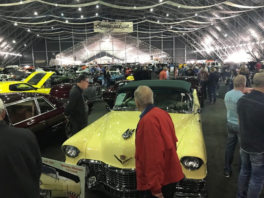 A car investor in a red jacket looking at a yellow vintage Cadillac with other bidders at a car auction