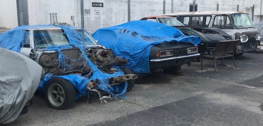 Silver Chevelle, black Camaro, green Corvette and silver Dodge van under blue tarps in a parking lot outside a restoration shop waiting on a car restoration project