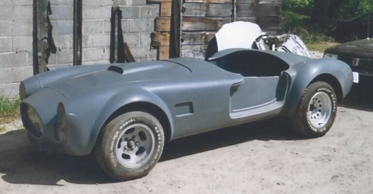 An unrestored gray Cobra sitting on dirt in front of a barn preparing for work using car restoration guidelines