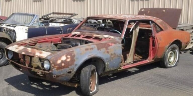 An orange Chevy Camaro without doors and trunk open sitting in front of a blue Camaro convertible in a parking lot waiting on a car restoration project