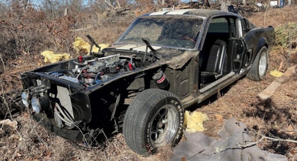 A black Ford Mustang GT500 without hood and doors as well as other missing parts in a field showing the classic car restoration risks