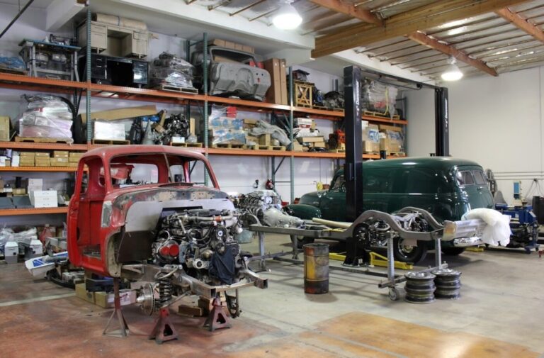 2 trucks; a red pickup and green panel truck and a silver frame inside a classic truck restoration specialty shop with parts stored on wall racks