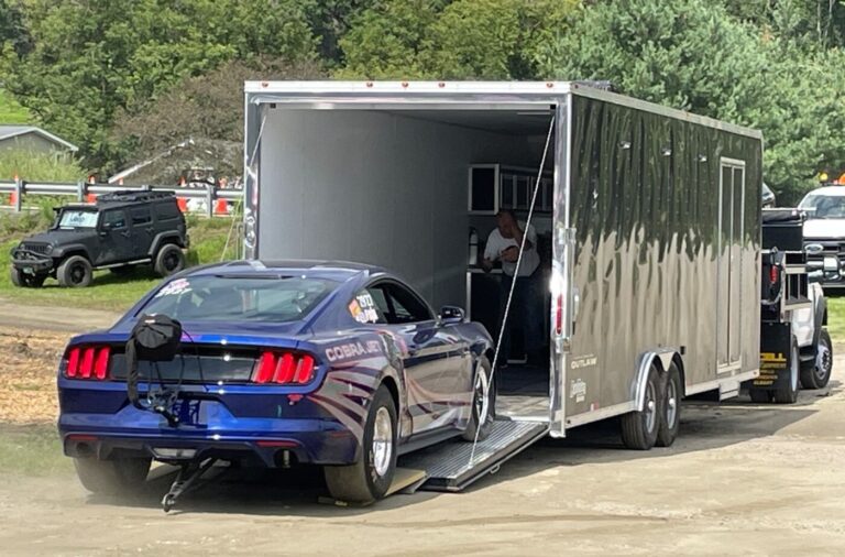 A modern blue Ford Mustang Cobra Jet race car driving onto an enclosed car trailer for classic car transporting