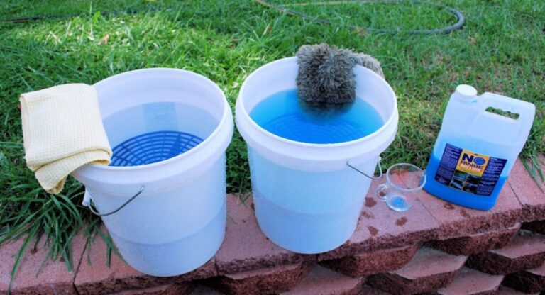 2 white buckets for classic car washing, one with clean water and a towel and the other with car wash and Mit along with car wash concentrate all sitting on a stone retaining wall
