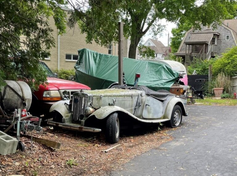 A gray vintage MG roadster parking next to a boat and red truck near a driveway needing inspection to determine if the car is not worth restoring