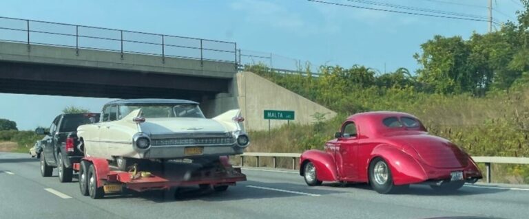 A white Cadillac on an open red car trailer being towed by a black pickup truck driving next to a red Willys cope on the highway