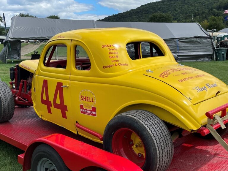 A yellow with red lettering dirt track race car on a red open flat bed car trailer parked at a car show with mountains in the background