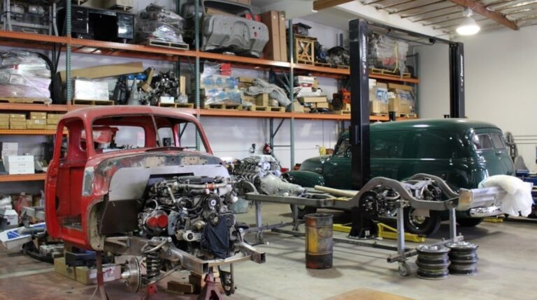 2 trucks iin a restoration shop, one being a red Chevy pickup and the other being a green Ford panel truck with parts in the background on wall shelves; it is important to know where parts come from to avoid car restoration shop scammers