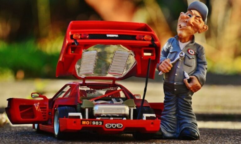 A cartoon with a mechanic in gray overalls holding a wrench and spark plug standing the rear of a toy red Ferrari with the bonnet open