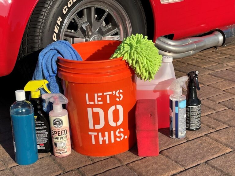 An orange bucket with white letters of Let's Do This and other cleaning supplies on a driveway in front of a red Cobra getting ready for car washing