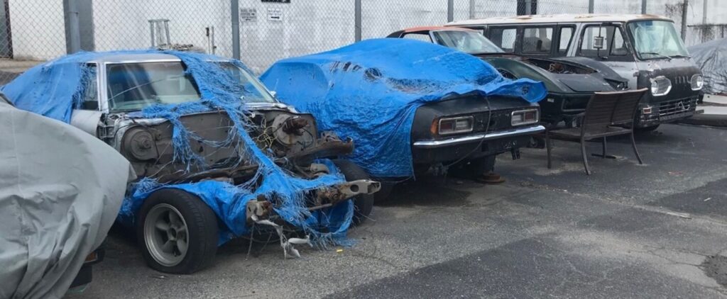 A Chevelle, Camaro, Corvette and Dodge van under blue tarps in a parking lot waiting on restoration as most people do not report restoration shop scams