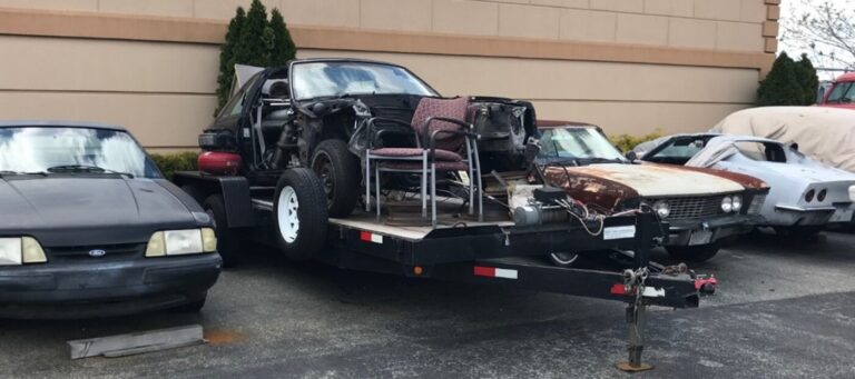 A black Ford Mustang, aa black disassembled Ford Mustang on a trailer, a gray Corvette, and a rusty Riviera parked outisde a restoration shop as the owners did not cut their car restoration losses