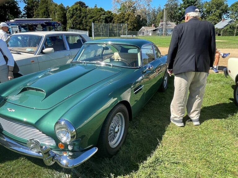 A man in a blue blazer doing a classic car pre-inspection on a green 60' Austen Healy i=on a grass field