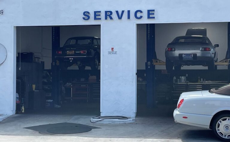 A view thru the garage doors of a white service station with an Alfa Romeo on the lift to the left and a silver Porsche 928 on the right and a Bentley parked at front prior to conducting a pre-purchase car inspection