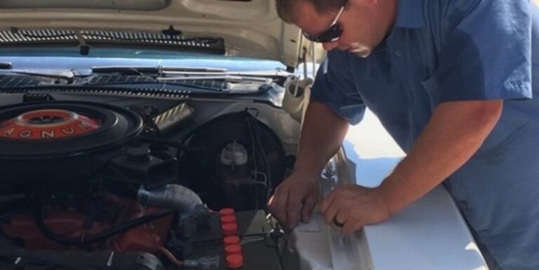 An inspector in blue shirt and sunglasses reviewing the information plate on a white Plymouth as part of the pre-purchase car inspection