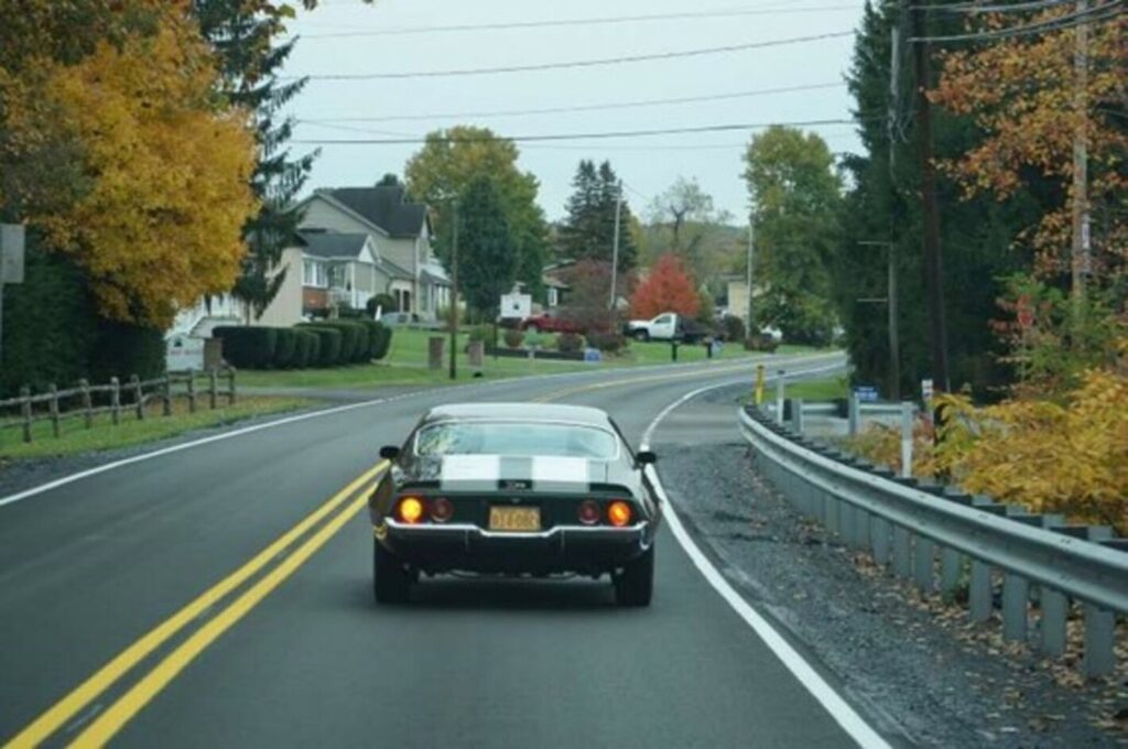 A 71 Chevy Camaro Z28 with white stripes driving on a 2-lane rural road with houses and guard rails showing on a twisting road why a car's weight distribution matters.