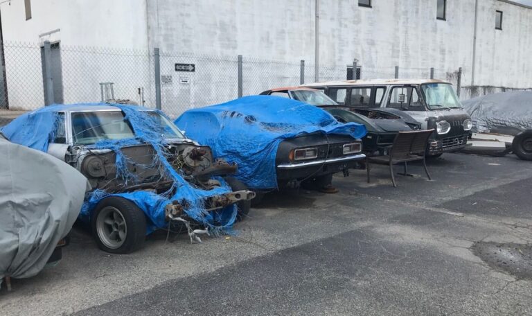 A group of old cars under blue tarps awaiting restoration in a parking lot as needing a written agreement is crucial in restoration success