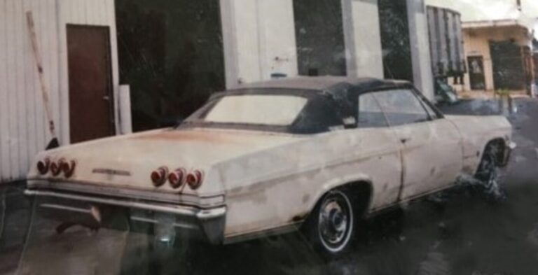 A rear right side view of a beige 1965 Chevy Impala convertible parked in front of a warehouse before starting the classic car DIY restoration