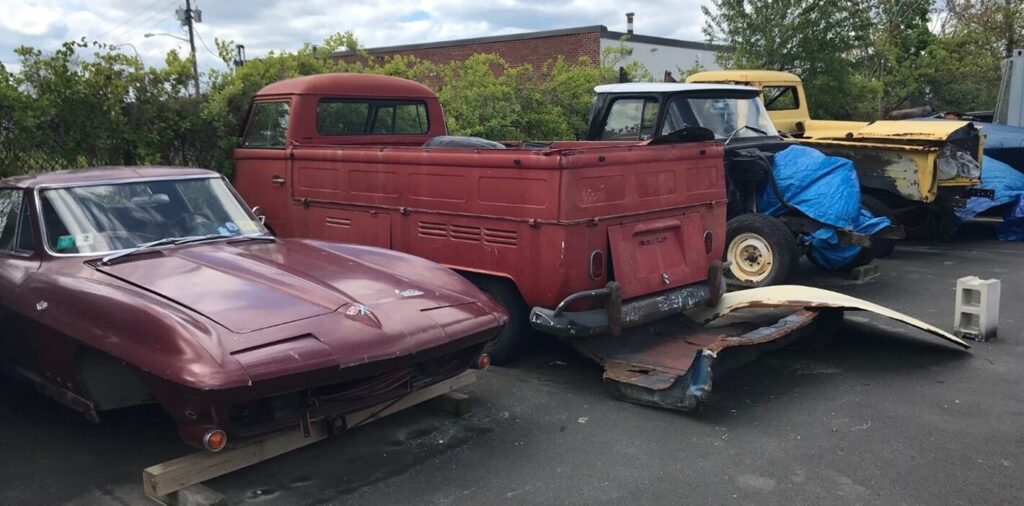 A burguundy Chevy Corvett on wood blocks parked next to a VW van as well as a yellow and black Chevy pickup trucks in a parking lot unrestored due to problematic car restoration contract cluases