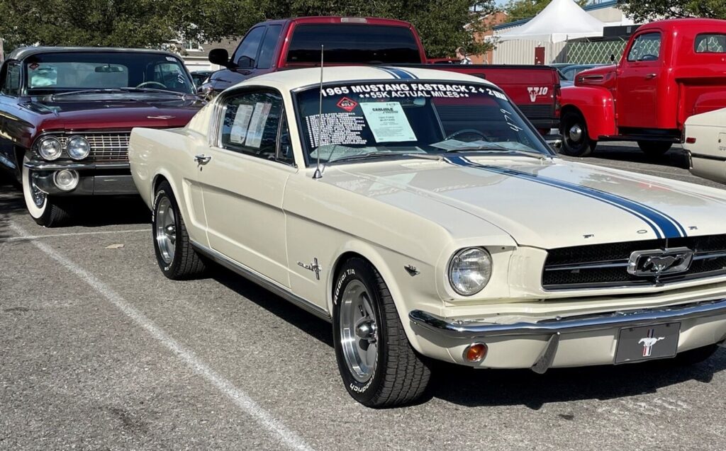 A white with blue stripes 1965 Ford Mustang sitting in an auction parking lot next to a red Cadillac deciding is that vintage car is worth the effort and expense