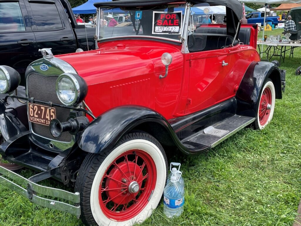 A red with black top Ford Model A convertible with a For Sale sign in the window on the grass at a car show while inspecting it to determine if the car is worth the effort and time to own