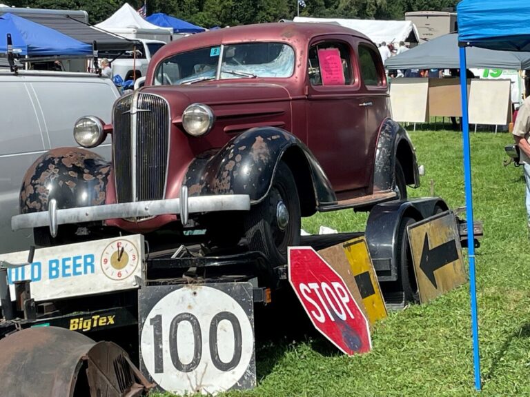 An unrestored 1940s red with black fenders Chevy sedan on a trailer surrounded by traffic signs on the grass at a car show