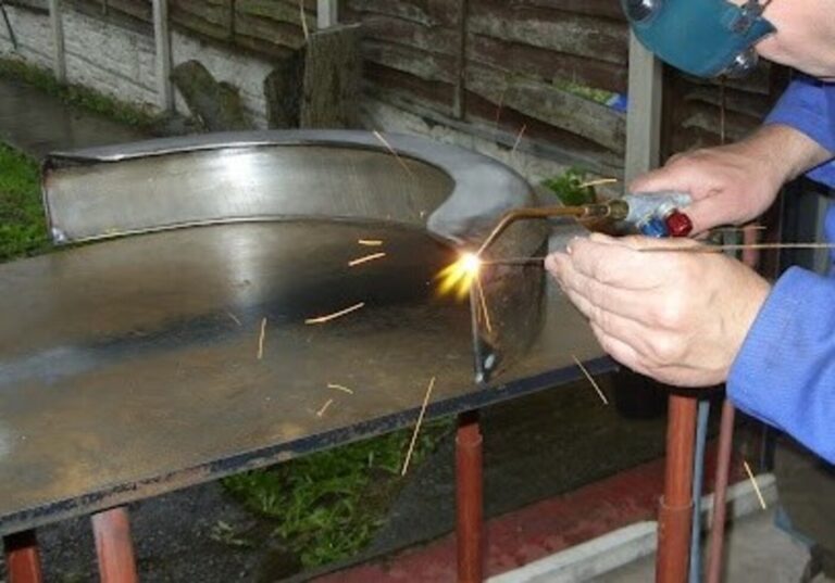 A mechanic in a blue shirt and eye shield working on welding repairs of a fender