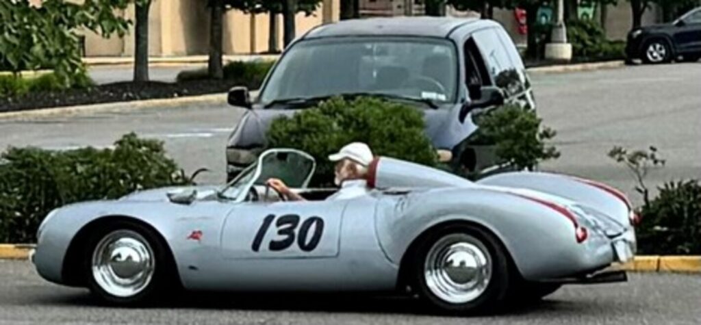 A man with white beard and hat driving a silver Porsche with the number "130" on the side door at a car show driving in extreme heat