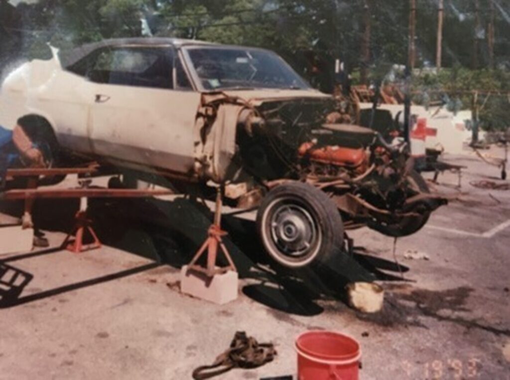 A beige 1965 Chevy Impala on jack stands wit hthe front clip removed and a mechanic working on the rear end with the essential tools needed for repairs