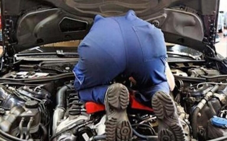 The view of the rear end of a mechanic in blue overalls and boots working in the engine compartment as part of a car restoration