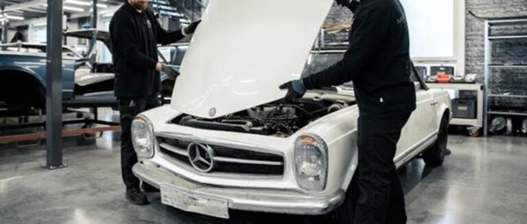 Mechanics in black overalls installing a hood on a white Mercedes Benz SL to repair accident damage requiring classic car insurance