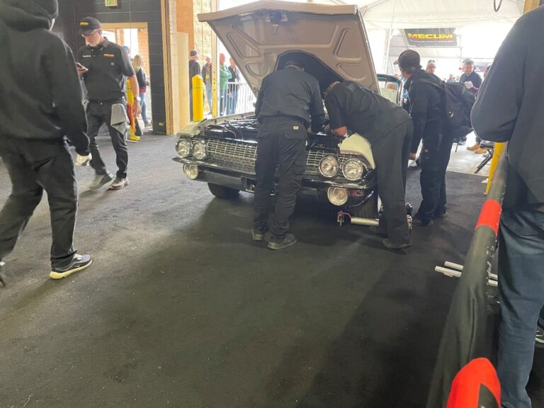 Mechanics working under the hood of a white Ford sedan at a car auction while peolpe look on