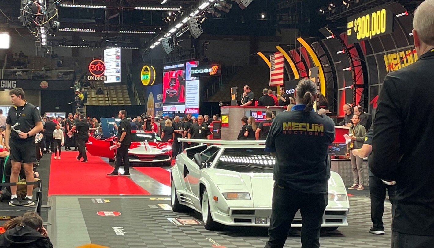 A white Lambo Countach followed by a red Ferrari crossing the auction block as bidders look on as they are classic car investing